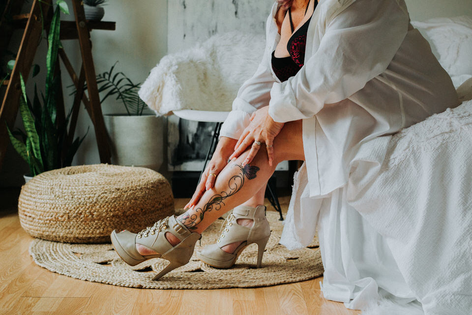 woman sitting on edge of bed leaning over to touch her tattooed leg wearing high heels, lingerie and a white shirt open over top