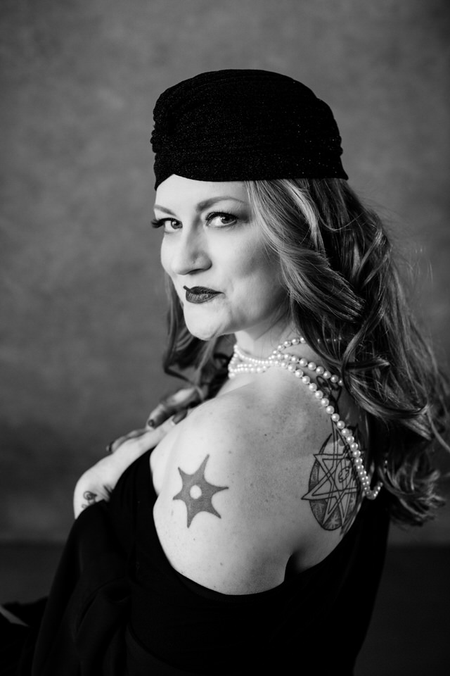 Black and white boudoir image of a bare shouldered, tattooed woman wearing pearls wrapped around her neck and a black hat