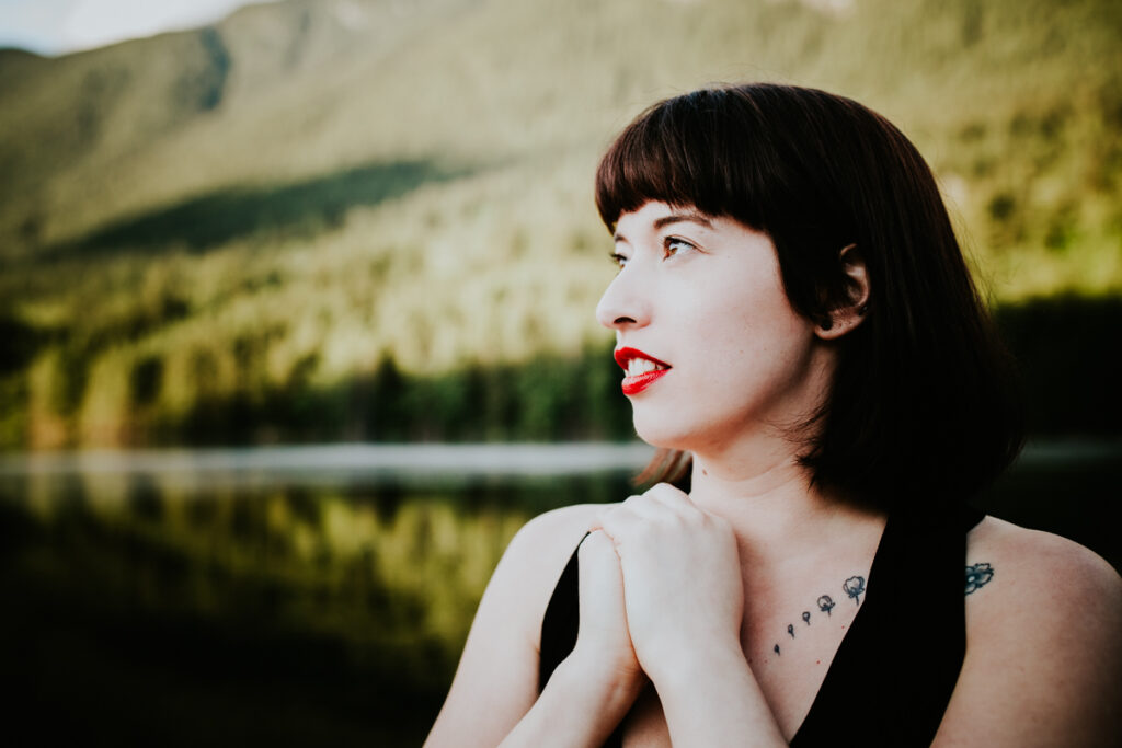 A tattooed woman with dark hair and red lips wearing a black tank looking to the distance with a lake and forested  mountain behind her