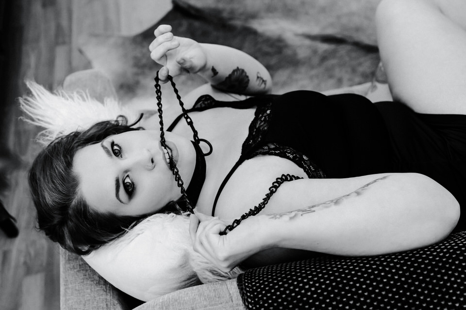 woman laying on her back wearing lingerie and a neck collar with a chain that she's holding in her hands and between her teeth in boudoir image