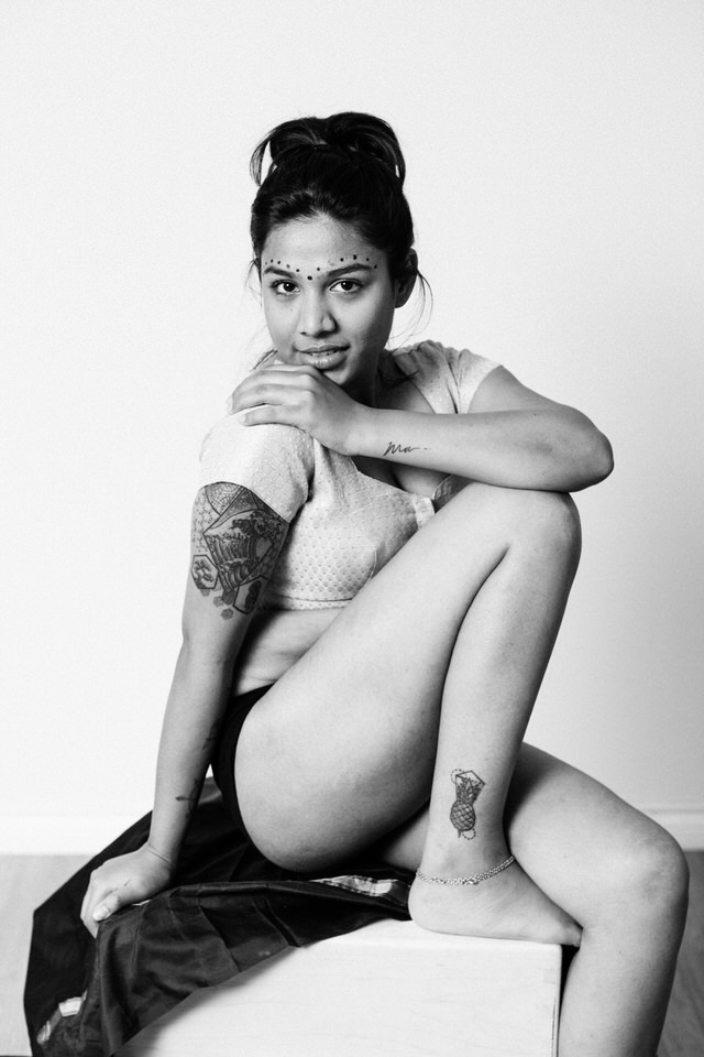 boudoir image of a tattooed woman sitting on a block with knee up and arm resting on her knee