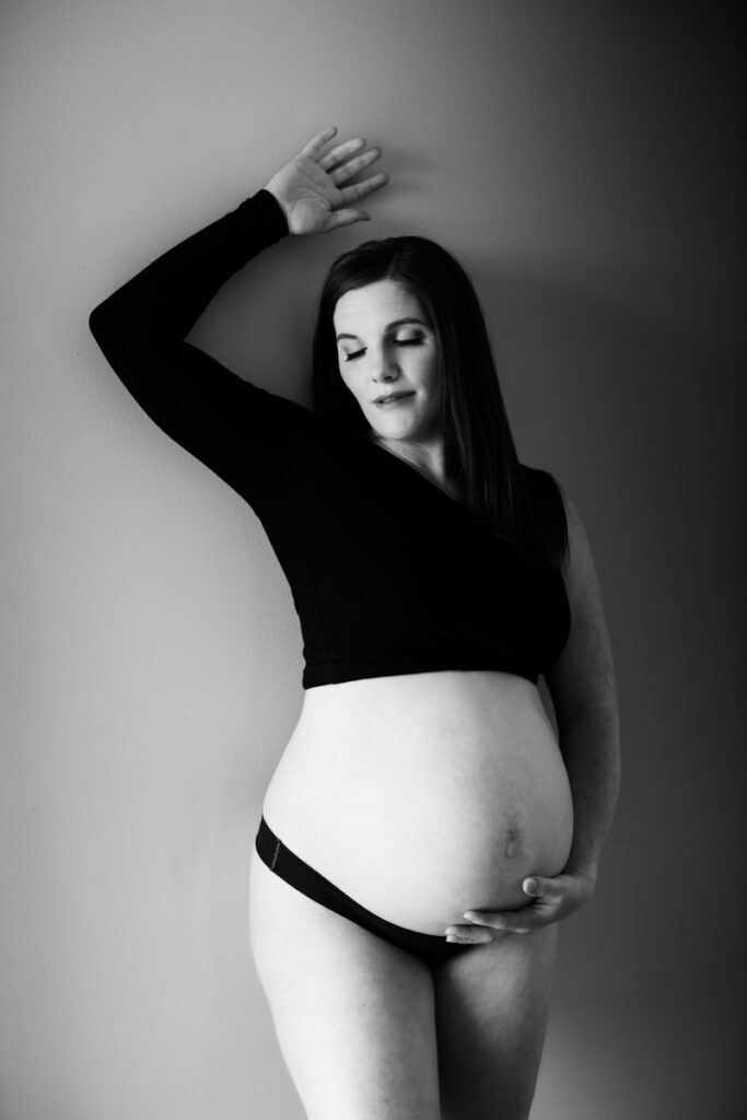 maternity boudoir photo of woman standing against the wall with one hand above her head and the other holding her bare belly