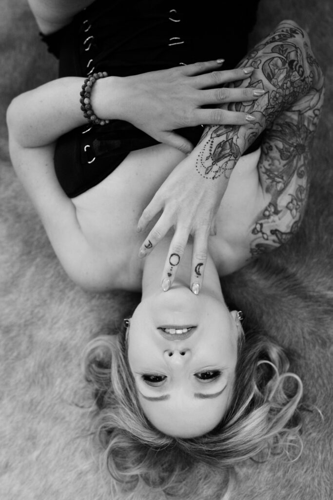 a fabulous woman over 40 laying on her back wearing a bustier, her right arm tattooed and right hand touching her chin and her left arm across her body