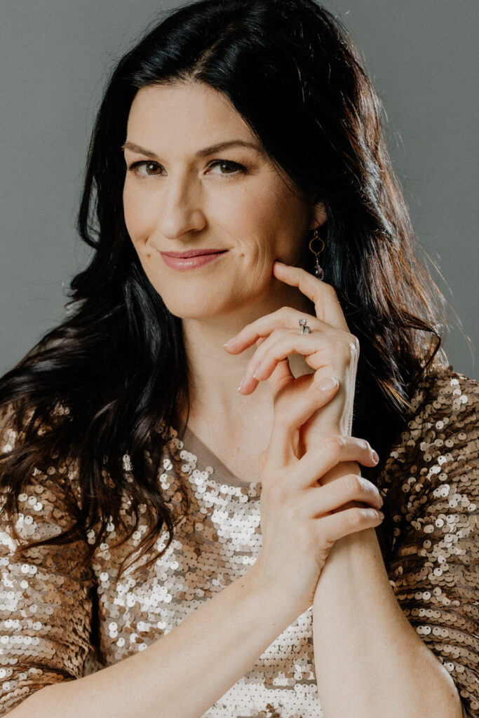 a luxe image of a woman in a gold sequin top smirking at the camera with her hands up at her face
