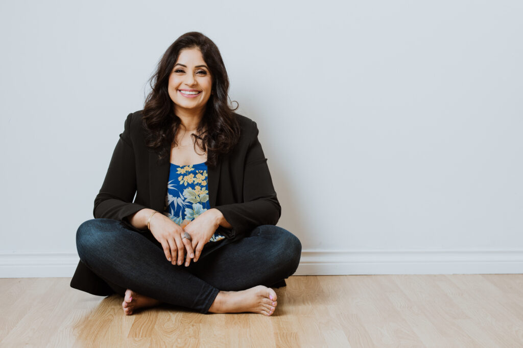 luxe personal branding portrait of a woman sitting crossed legged against a bare white wall, she's smiling wearing a blazer and jeans