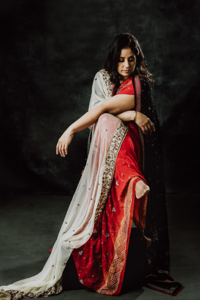 a luxe intimate beauty portrait of a woman sitting on a stool wearing a sari draped down to the floor, her leg crossed over the other and her arms draped criss-crossed across her body