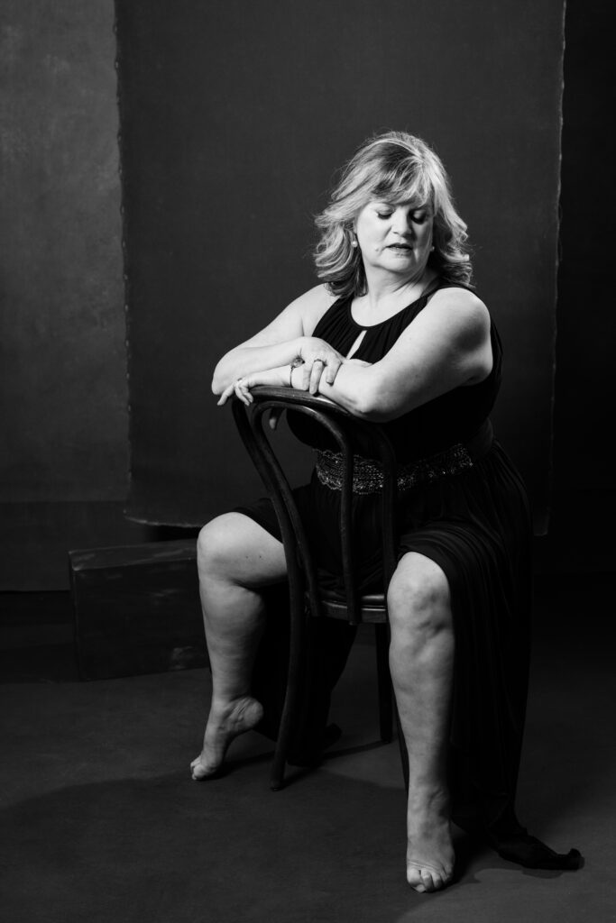 boudoir image of a woman over 40 sitting backwards on a chair wearing a black dress