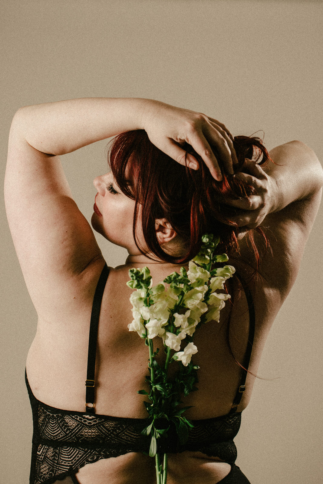 Boudoir photo of woman's back reaching up to her head and holding flowers hanging down her back