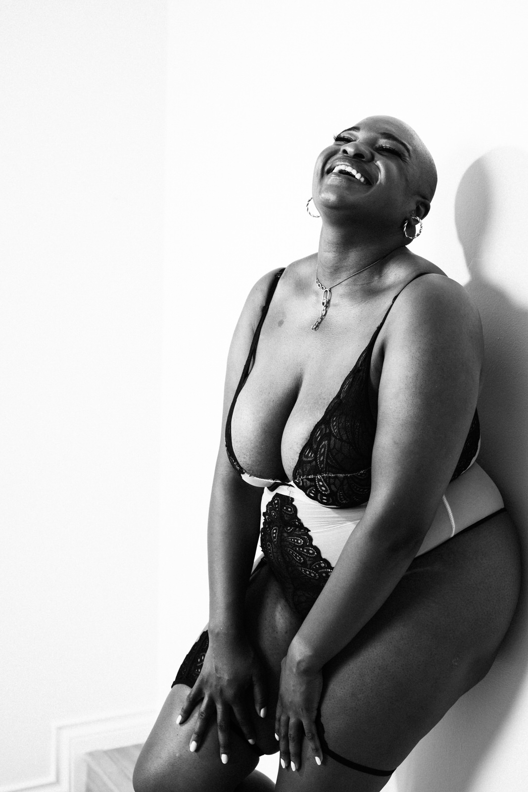vancouver boudoir photographer image of plus size black woman with alopecia wearing lingerie and leaning on wall with head tilted back laughing