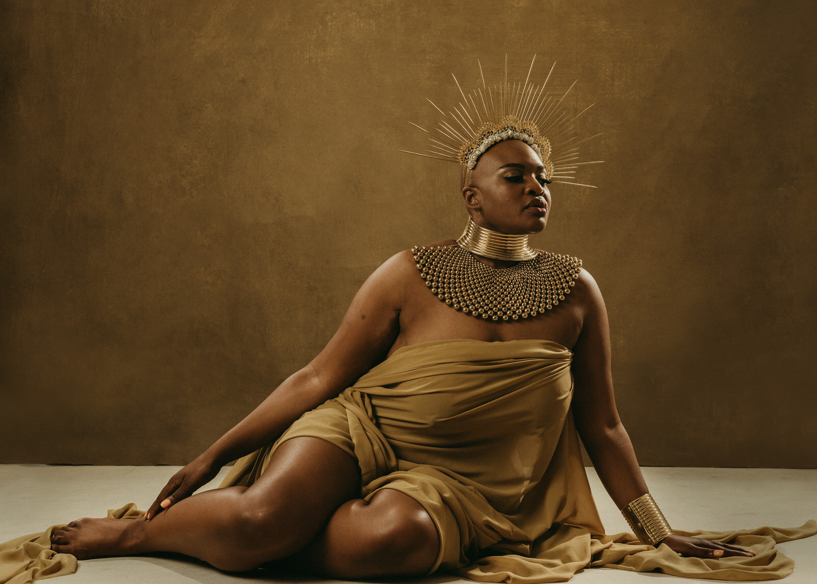boudoir photo of plus size black woman with alopecia sitting on floor draped in fabric wearing neck piece and sun crown
